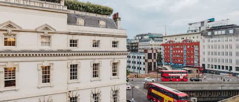 A city street with a red double-decker bus, buildings with classical architecture, and a clear sky.