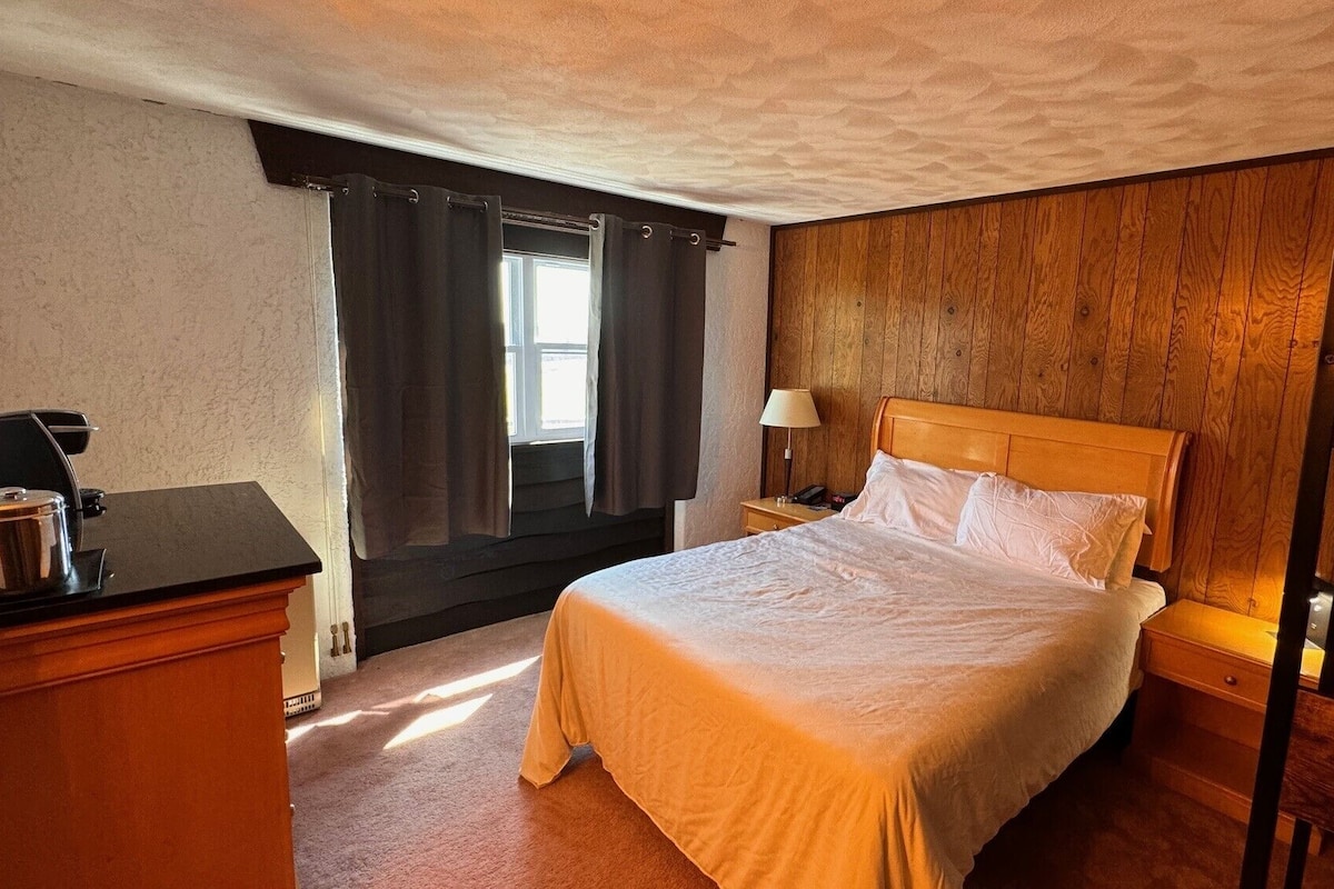 A hotel room with a wooden headboard, a bed with white linens, a nightstand with a lamp, a desk with a coffee maker, and a window with curtains.