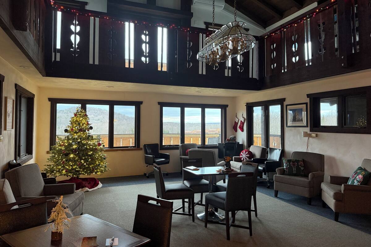 A spacious living room with a large Christmas tree, multiple windows, and a high ceiling with exposed beams.