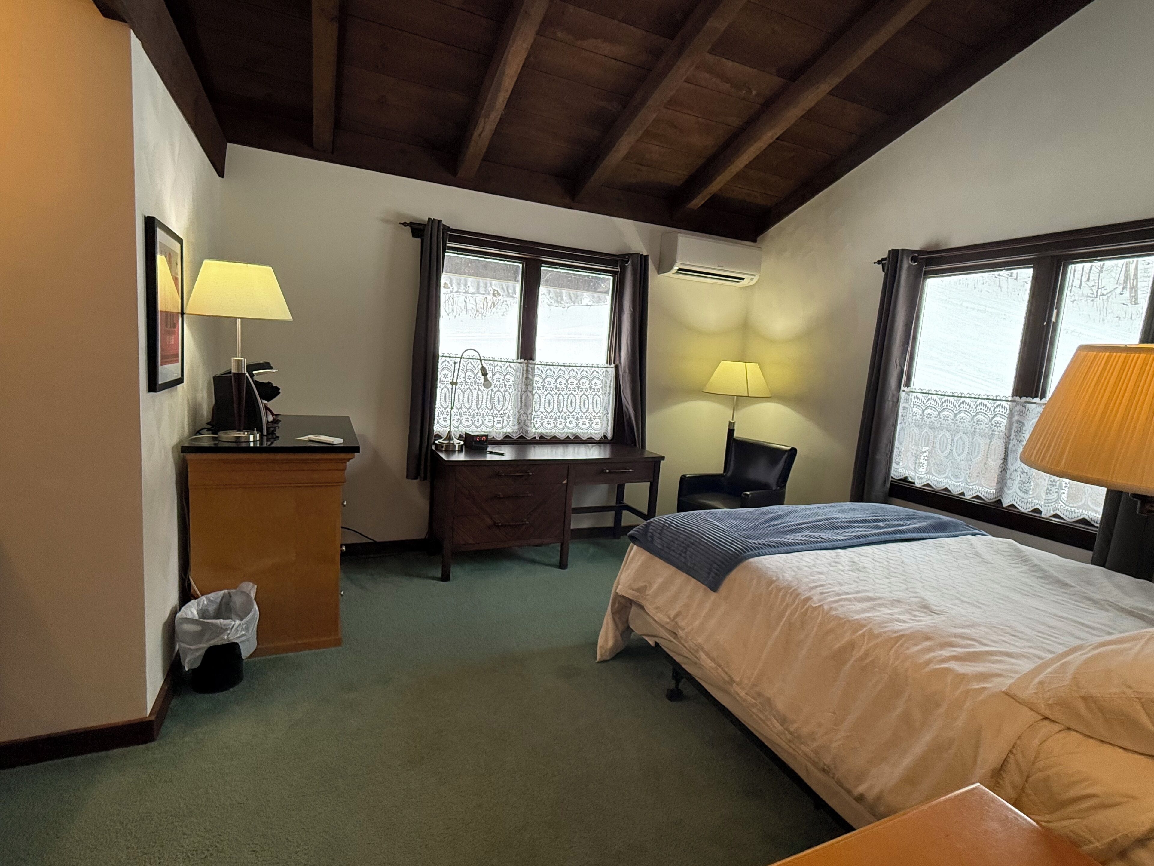 Une chambre d’hôtel avec un lit, un bureau, une chaise et deux fenêtres.