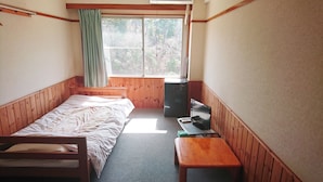 A small, single-room cabin with a bed, a desk, a chair, a small table, a refrigerator, and a window with a view of trees. - Hotel Sunrise (Onagawa)