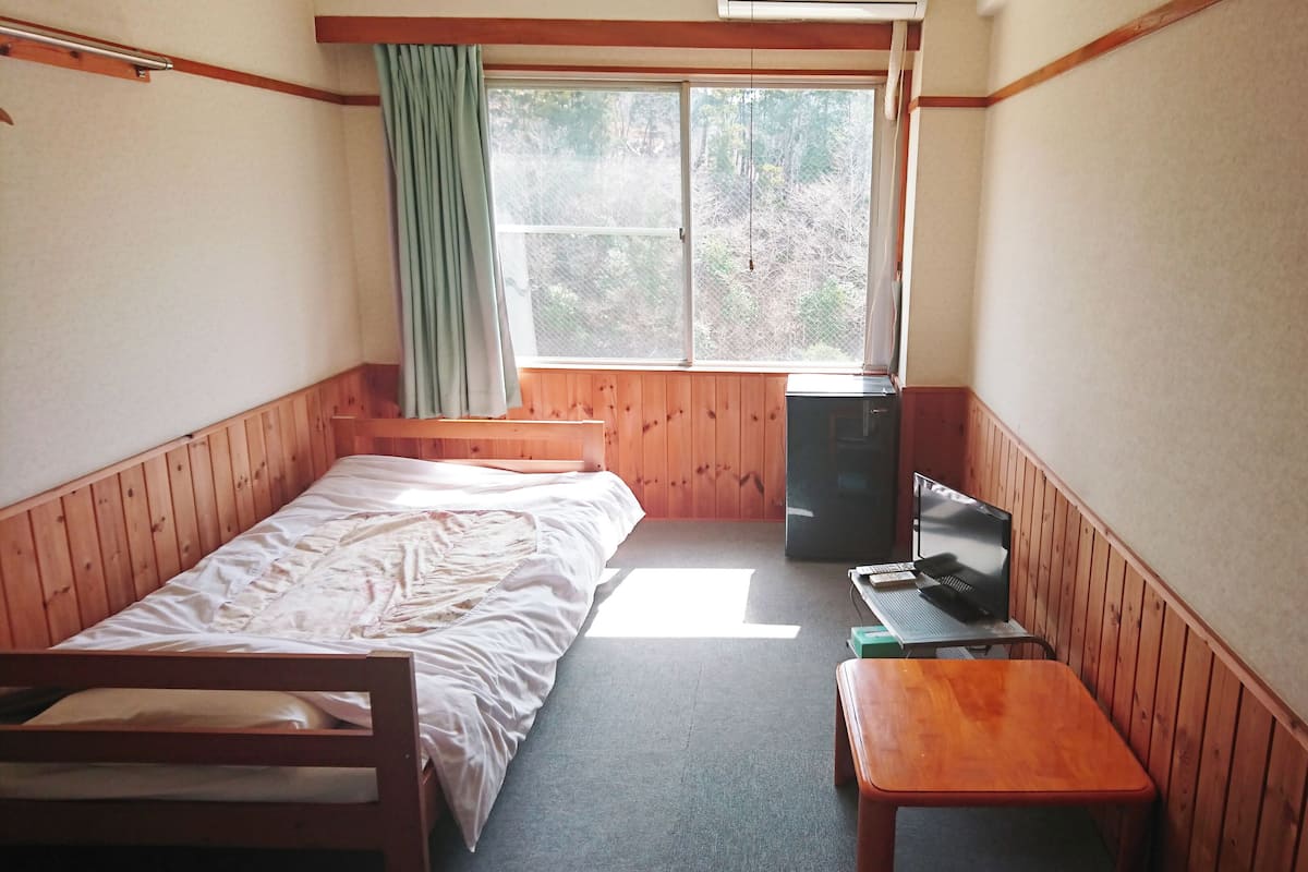 A small, single-room cabin with a bed, a desk, a chair, a small table, a refrigerator, and a window with a view of trees.