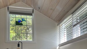 A bedroom with a bed, a bedside table with a lamp, a window with blinds, and a view of greenery outside. - Cosy bespoke Cottage in Olinda-central location (Olinda)
