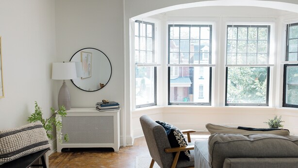 A living room with a sofa, a chair, a small table with a lamp, and a radiator. There are windows with blinds and a view of buildings outside. - Walking distance to Wellspan York Hospital, York College. Comfortably sleeps 8. (York)