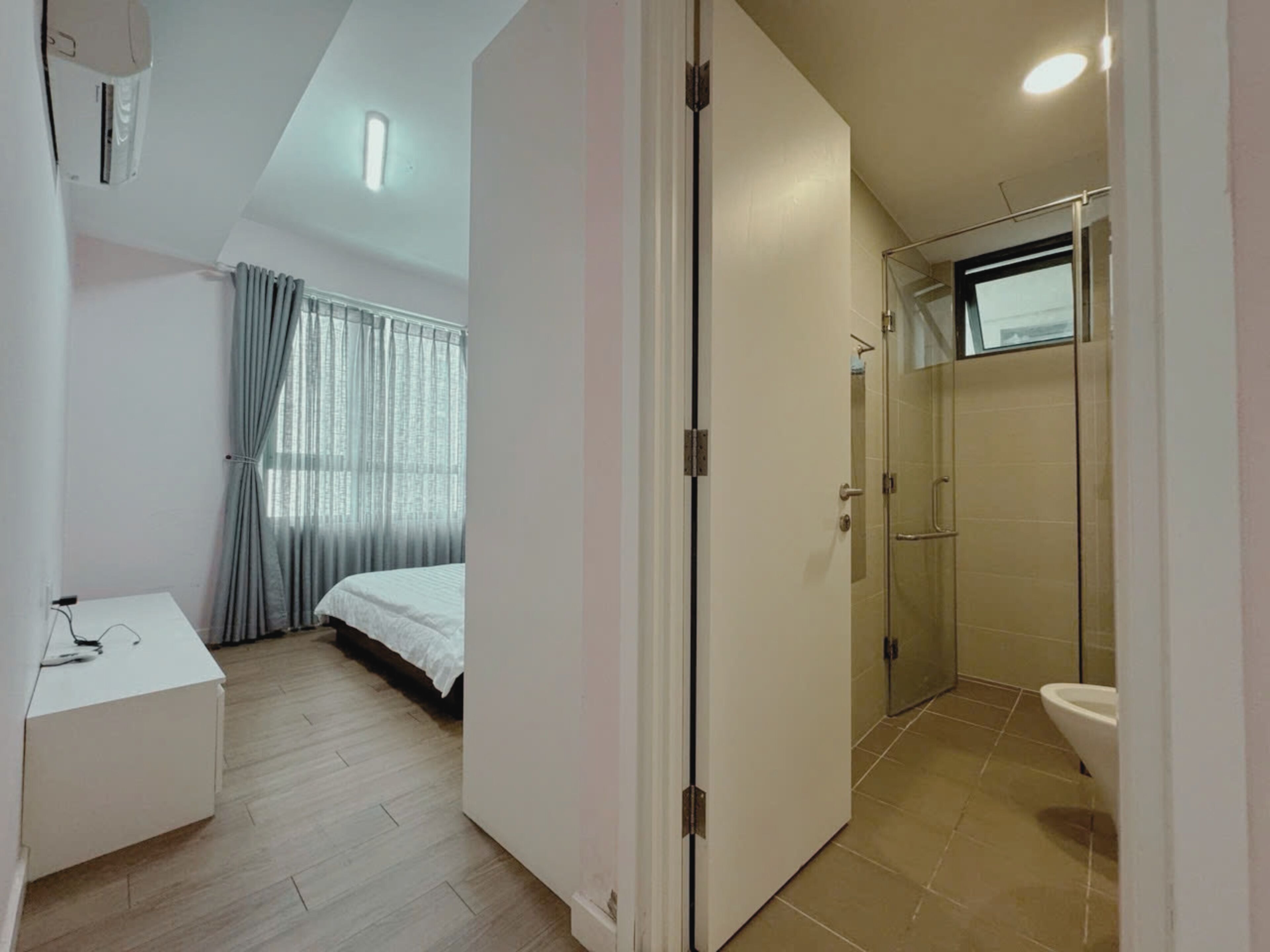 A bedroom with a bed, a bedside table, a window with curtains, and a bathroom with a shower and toilet.