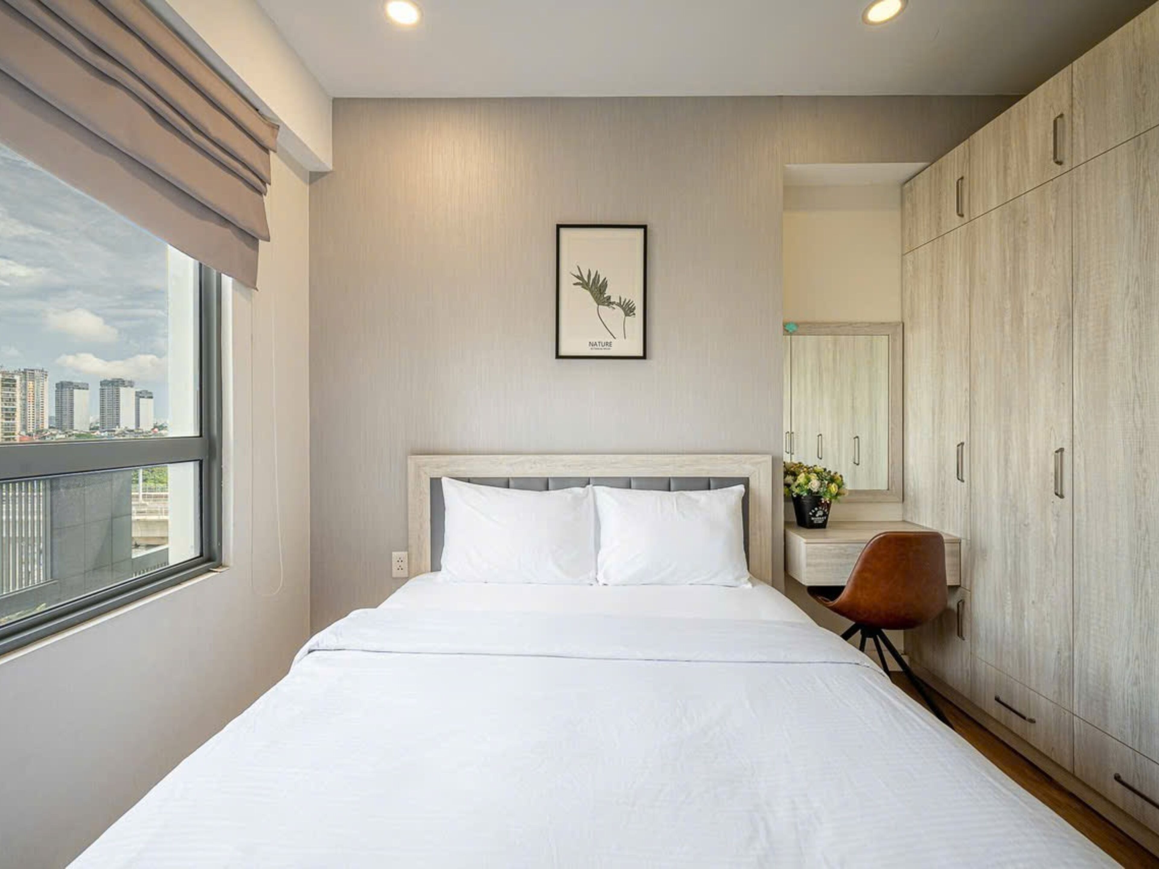 A modern hotel room with a large bed, a city view, a framed nature picture, and a wooden wardrobe.