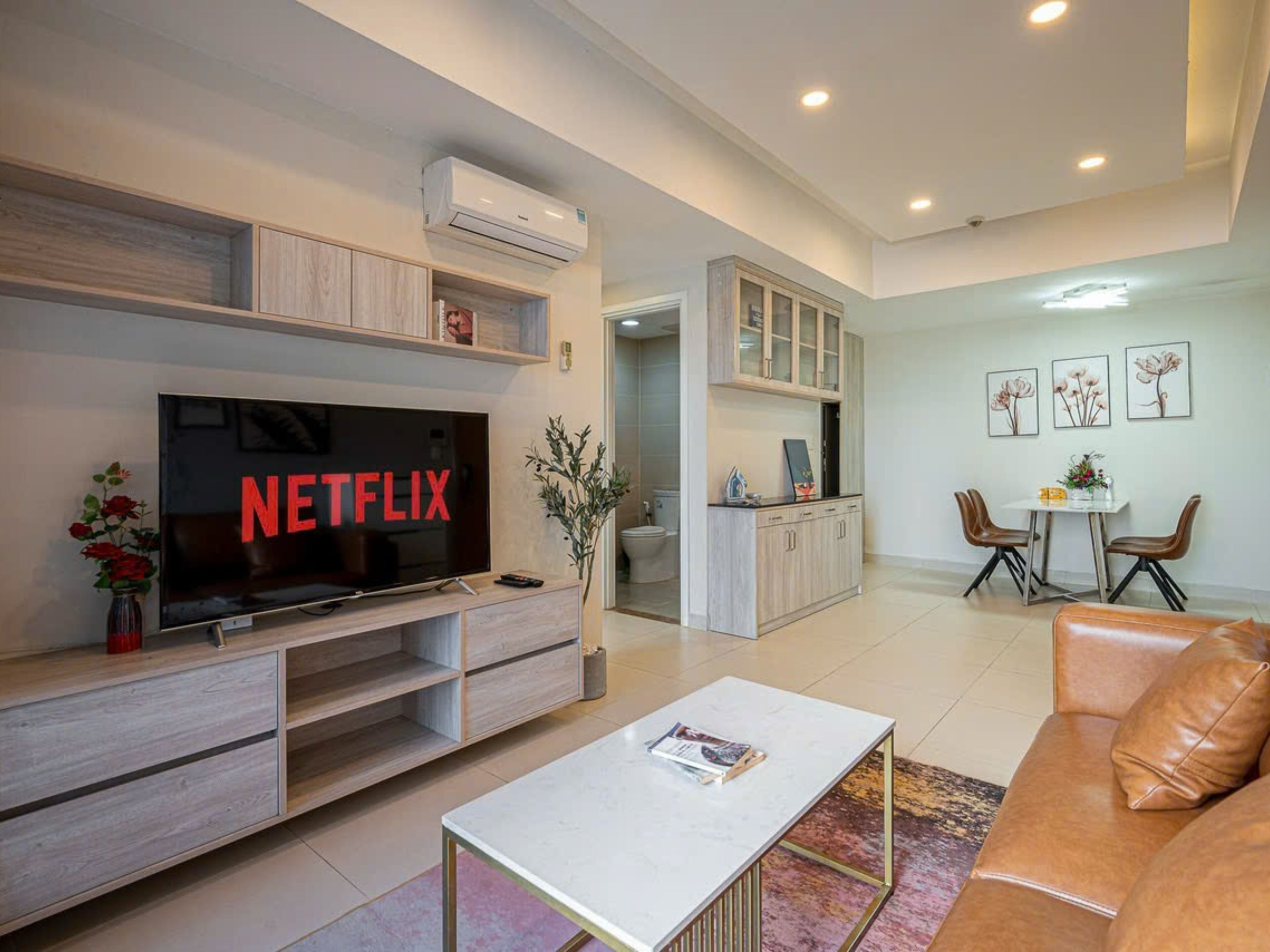 Un salon moderne doté d’un téléviseur à écran plat affichant le logo « NETFLIX », d’un coin repas avec une table et des chaises, et d’une kitchenette équipée de placards et d’un plan de travail.