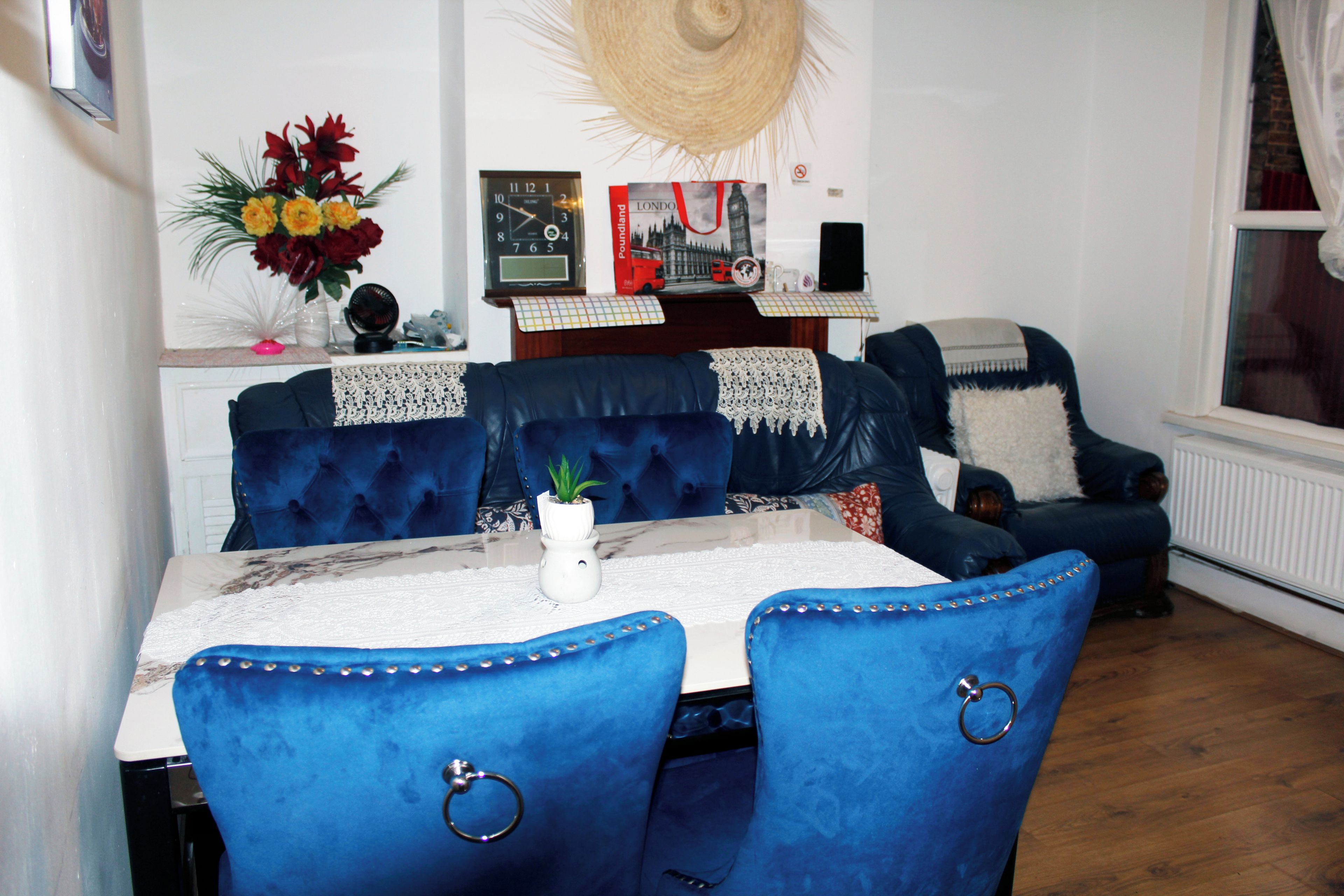 A dining area with a table set for four, blue upholstered chairs, a decorative vase with flowers, a wall clock, and a poster of London.