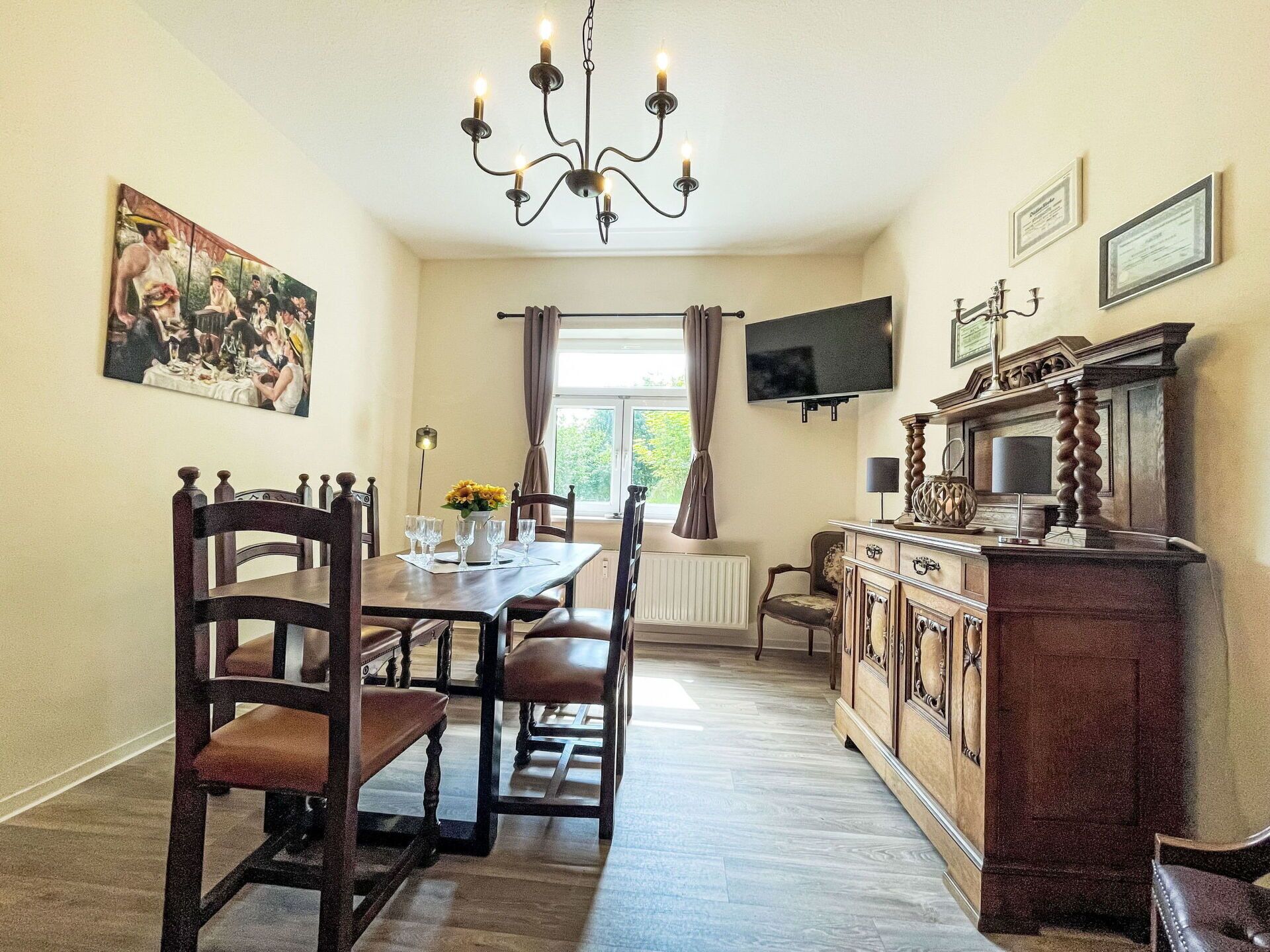 Ein Essbereich mit einer Kronleuchter, Holzsesseln, einem Tisch mit Blumenvase und einem Fernseher an der Wand.