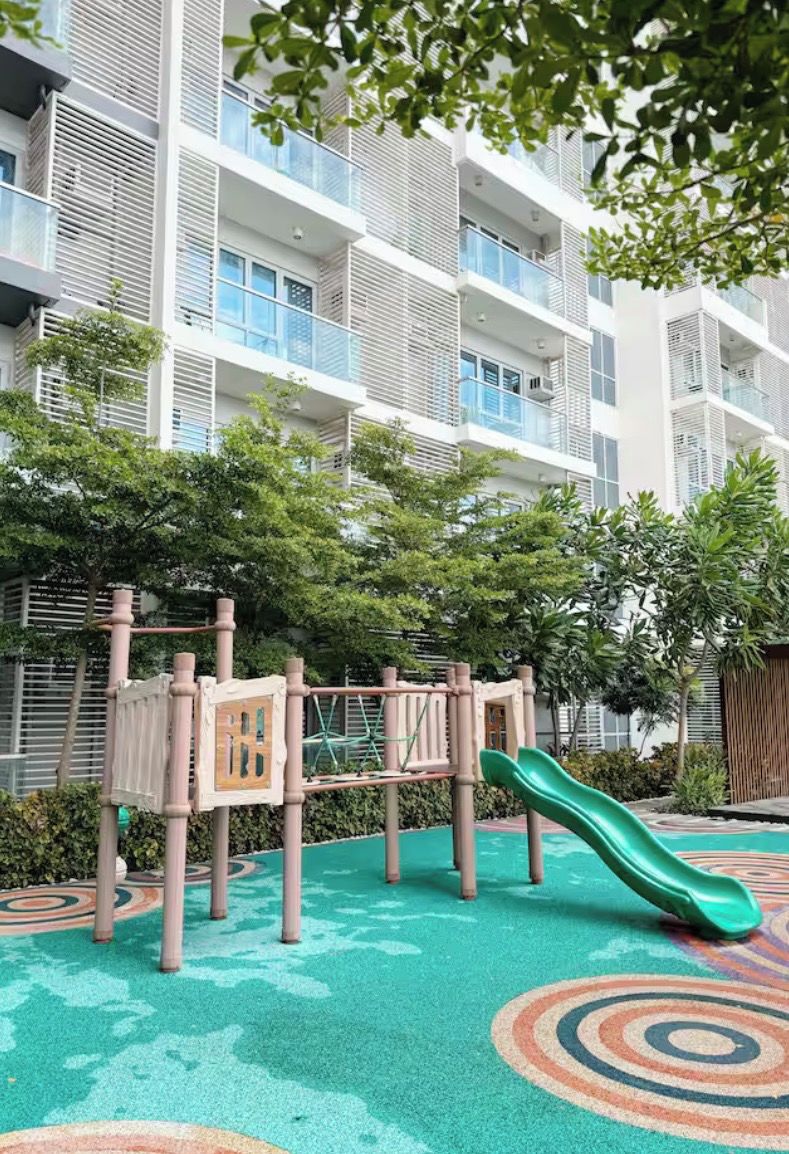 A playground with a slide and climbing structures, surrounded by a multi-story building with balconies and greenery.