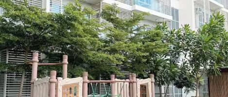 A playground with a slide and climbing structures, surrounded by a multi-story building with balconies and greenery.