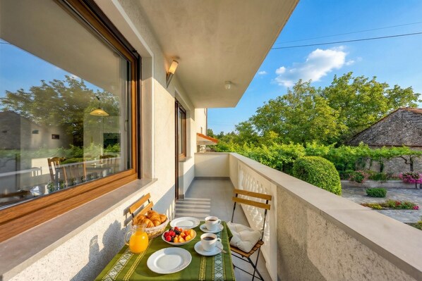 Un balcon aménagé pour le petit-déjeuner, avec des croissants, des fruits et du café, donnant sur un jardin où se trouve un bâtiment en pierre.