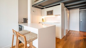 Een moderne keuken met witte kasten, een ingebouwde magnetron en een klein eiland met een spoelbak. Er staan twee houten stoelen met geweven zittingen.