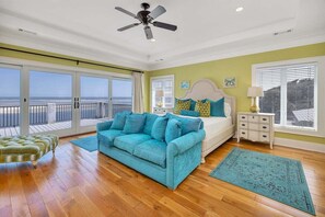 A spacious room with a large window, a ceiling fan, a bed, a blue sofa, and a small table. - Oceanfront Home Offers Breath Taking Views & Sunrise (Saint Simons Island)