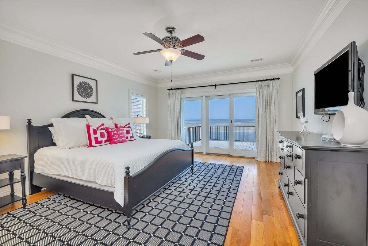 A bedroom with a bed, a ceiling fan, a TV, a dresser, and a view of the ocean through large windows.