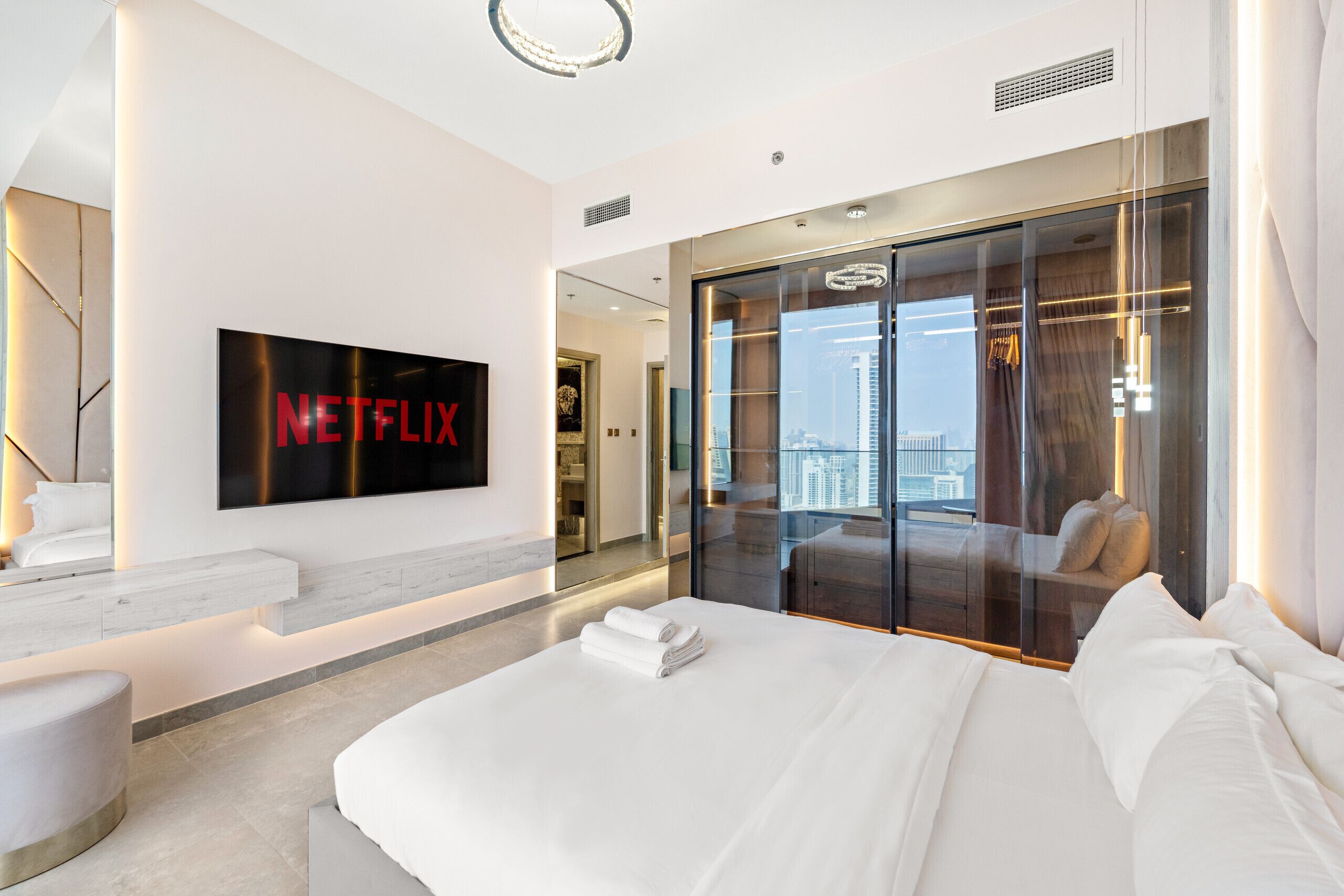A modern hotel room with a large bed, a flat-screen TV mounted on the wall, a walk-in closet with a view, and a cityscape visible through the windows.