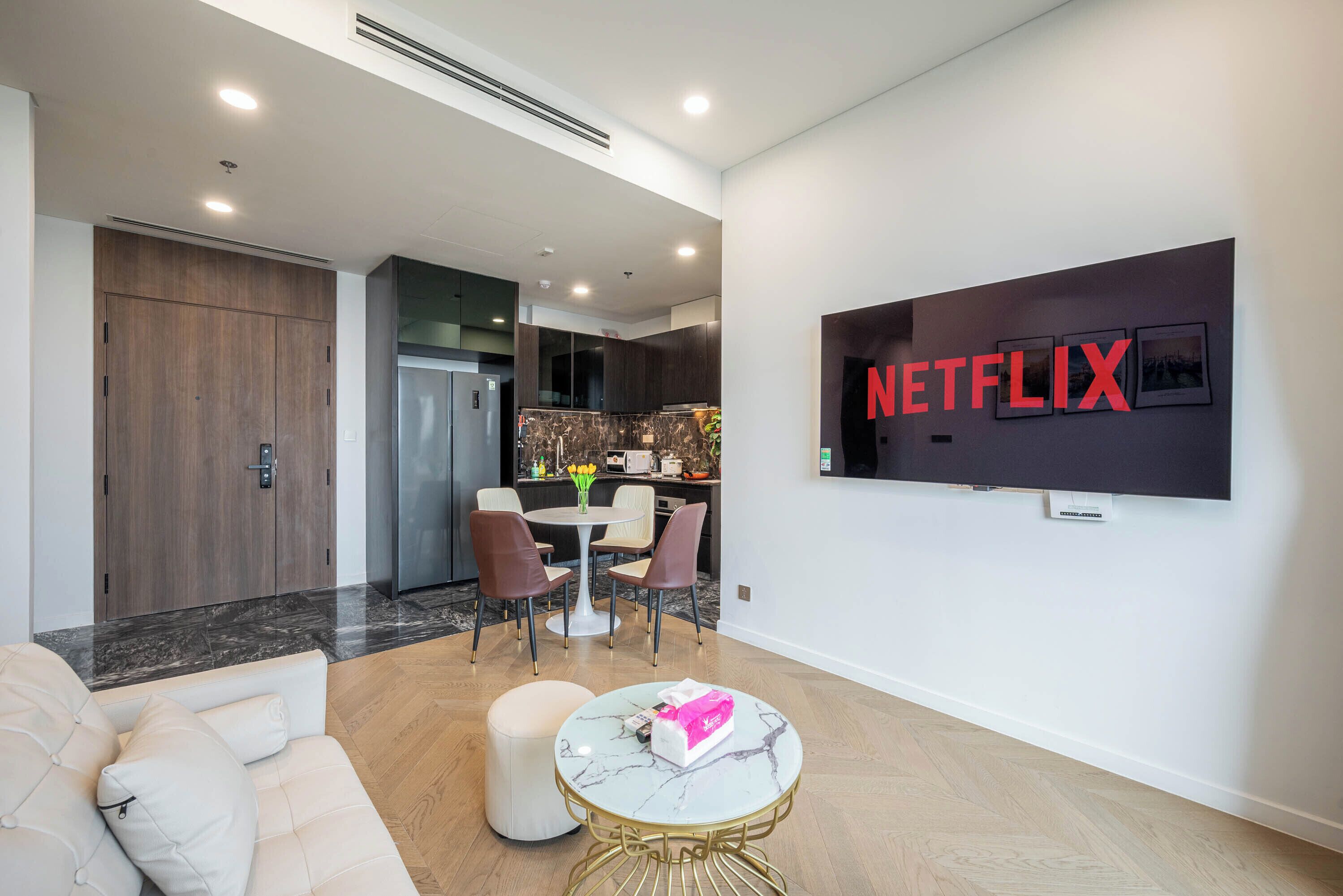 A modern living room with a sofa, ottoman, and a coffee table. There is a dining area with chairs and a table, and a kitchen in the background. A TV mounted on the wall displays the Netflix logo.