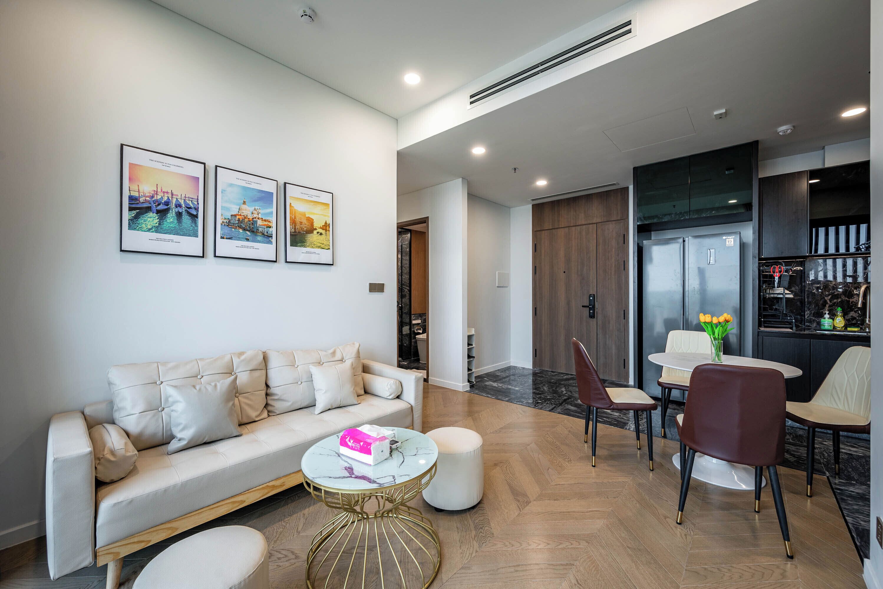 A modern living area with a sofa, a round table, and a dining set. There are framed pictures on the wall and a kitchen area in the background.