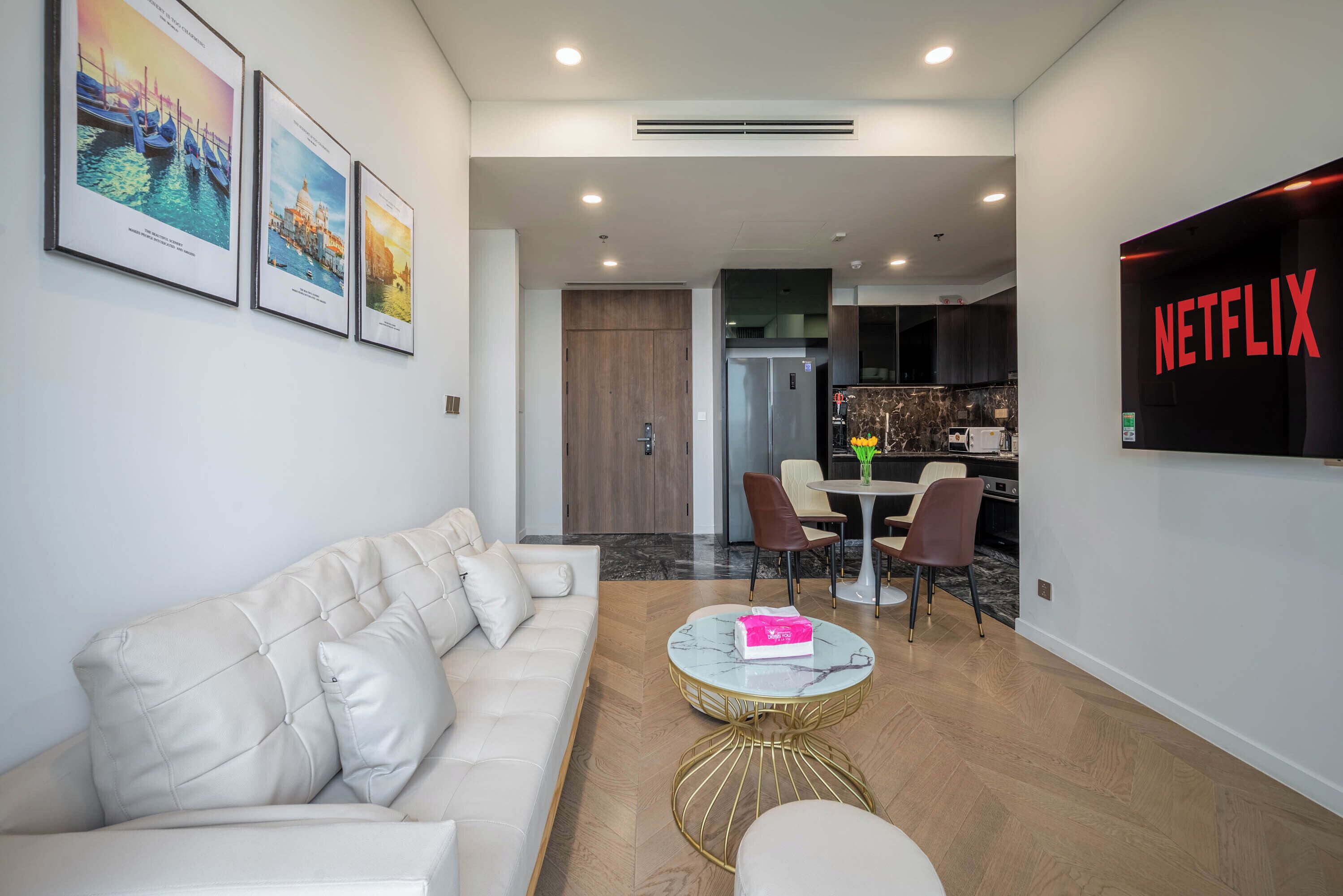 A modern living room with a white sofa, a round coffee table, and framed cityscape pictures on the wall. A kitchen area with a dining table and chairs is visible in the background.