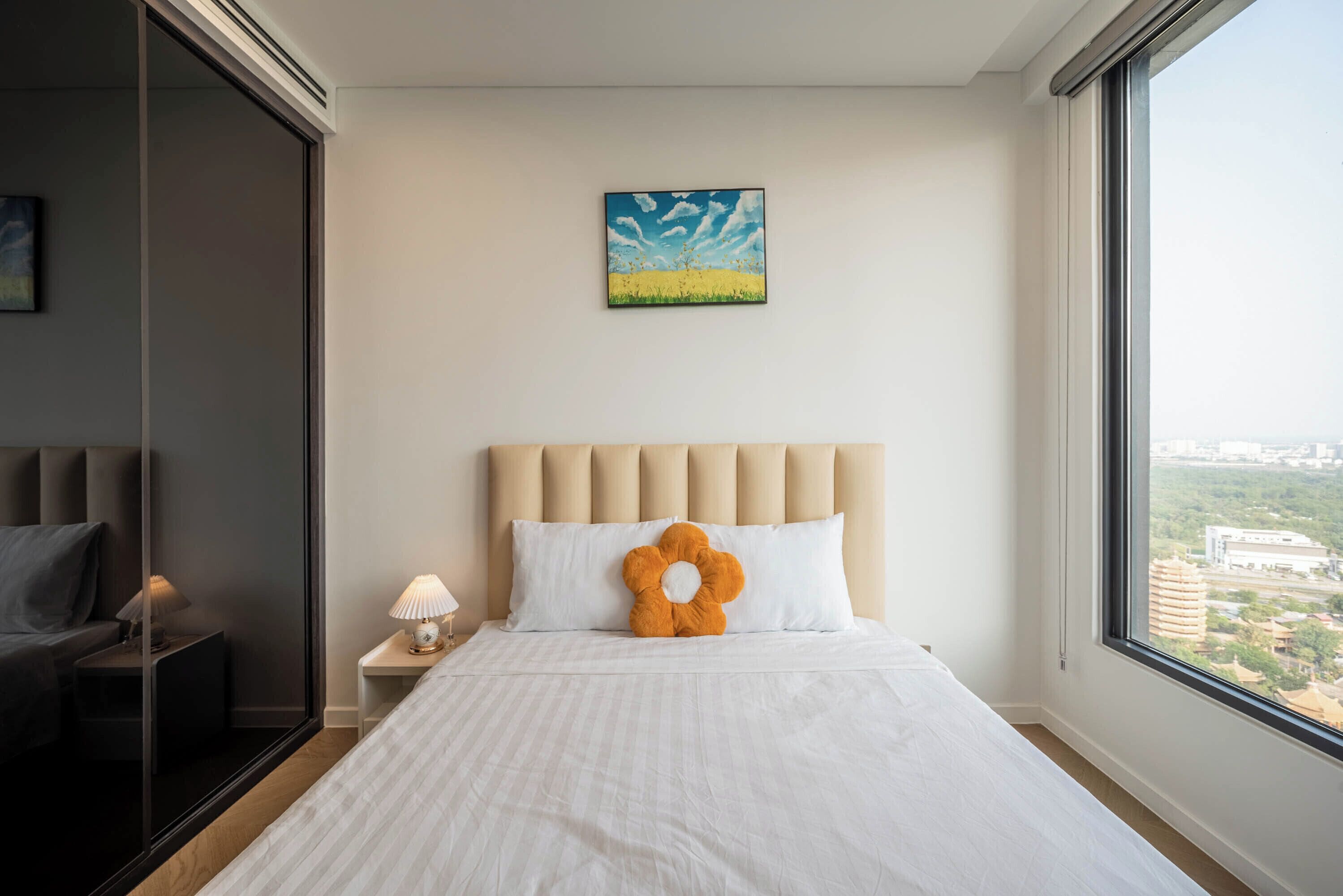 A hotel room with a large bed, a headboard, a nightstand, a lamp, a window with a city view, and a painting on the wall.