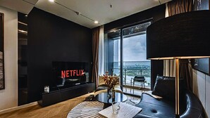 A modern living room with a large TV displaying 'NETFLIX', a sofa, a coffee table, and a view of the city through large windows. - The Lumiere Luxury Citypoint ANGIA (Ho Chi Minh City)
