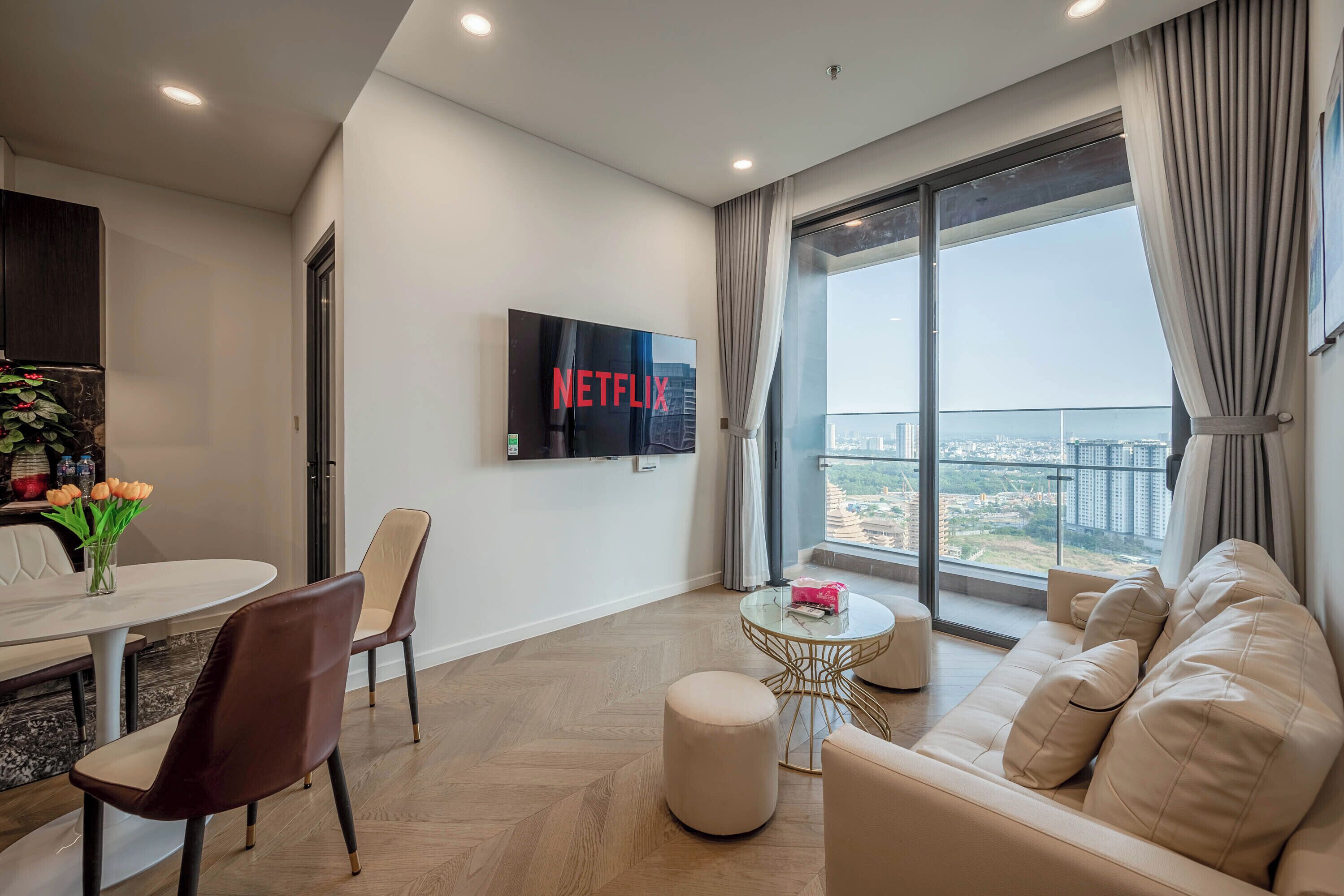 Ein modernes Wohnzimmer mit großem Wand-Fernseher, einer Couch, einem Esstisch mit Stühlen und Blick auf die Stadt durch große Fenster.