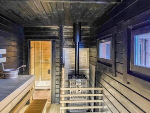 Interior de una cabaña de madera con chimenea central, un banco de madera y una ventana en la pared derecha.