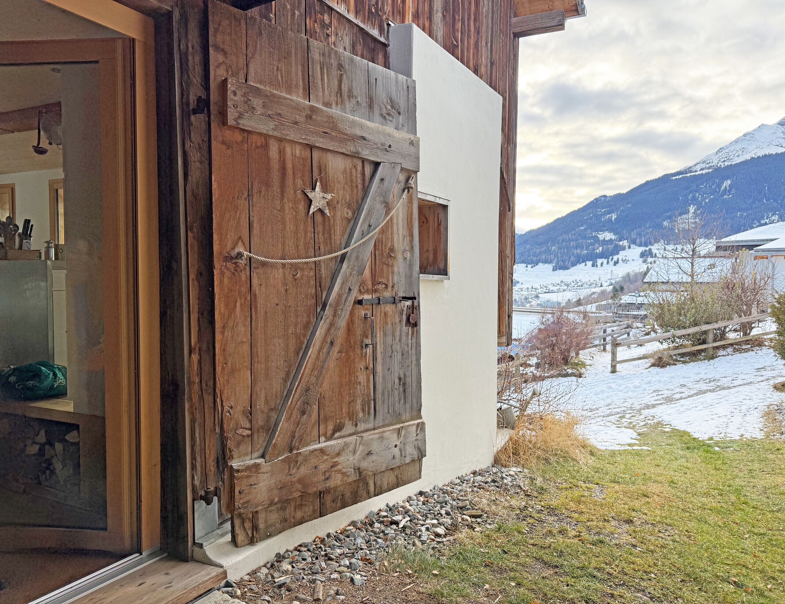 Una puerta de madera rústica con adorno de estrella, una cadena metálica y vista a un paisaje nevado con montañas al fondo.