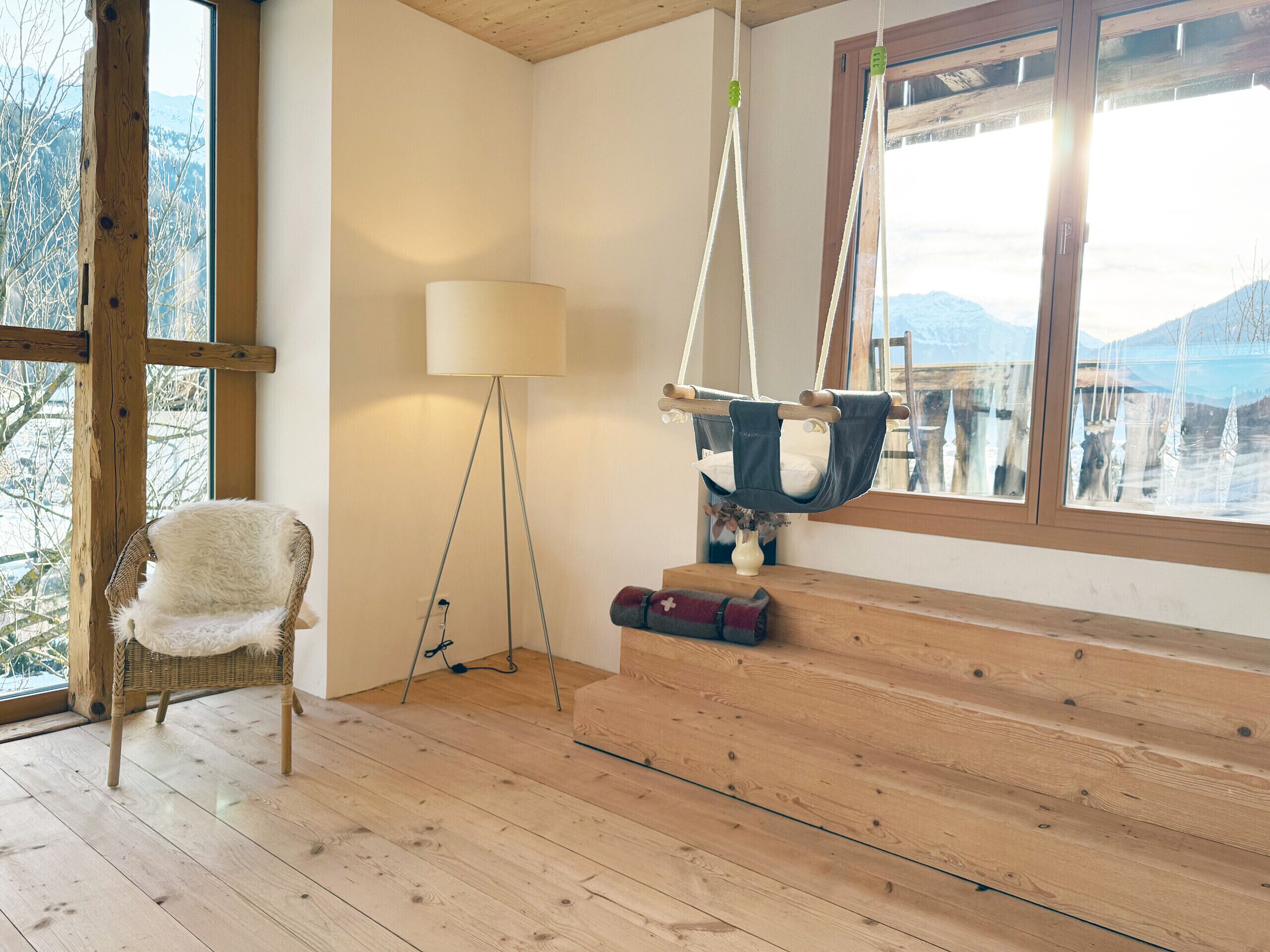 Une pièce chaleureuse avec un plancher en bois, un fauteuil en osier, un fauteuil suspendu et une vue sur les montagnes par une grande fenêtre.