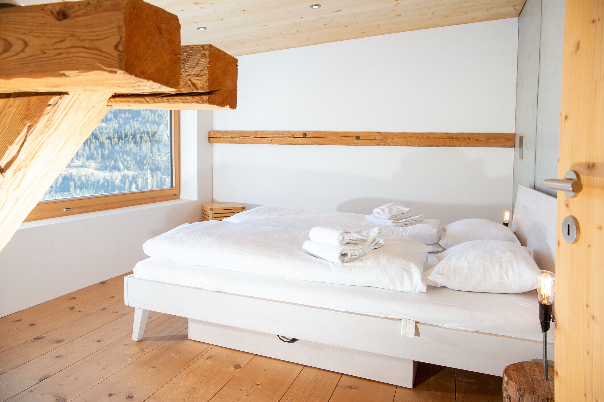 A bedroom with a wooden ceiling, a large window with a view of a forest, a white bed with neatly folded towels, and a wooden nightstand.