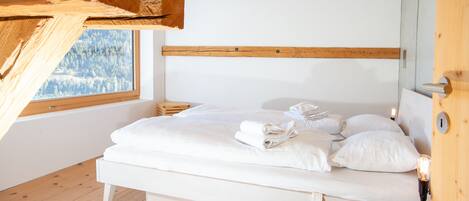 A bedroom with a wooden ceiling, a large window with a view of a forest, a white bed with neatly folded towels, and a wooden nightstand.