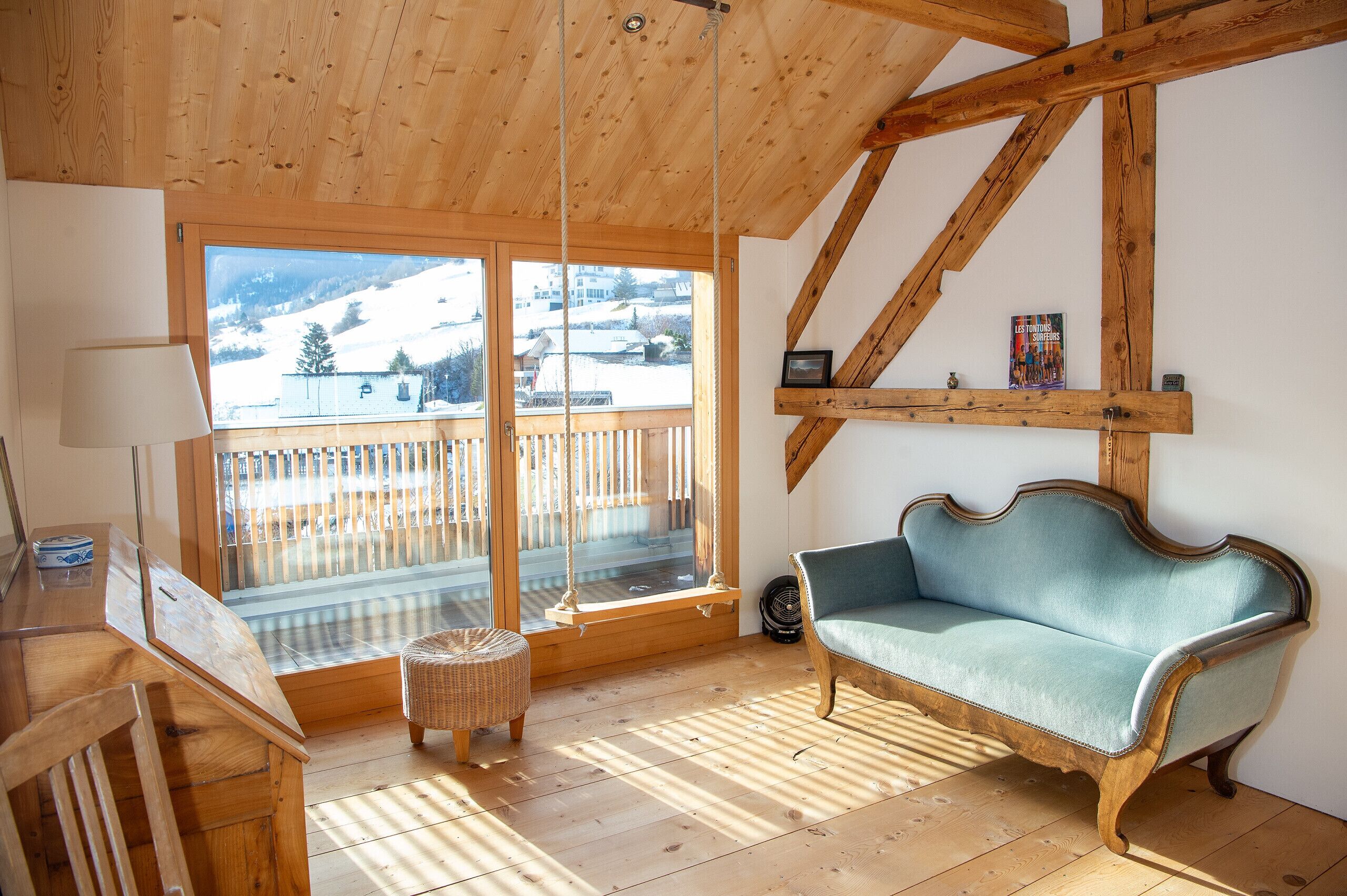 A cozy room with a wooden ceiling, a large window with a view of a snowy landscape, a blue sofa, and a wooden desk with a lamp.