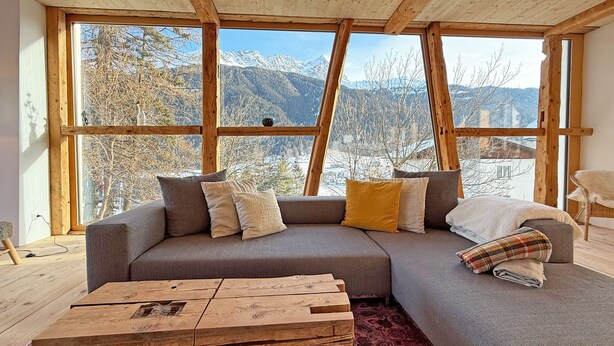 A modern living room with a large window, a gray sofa, and a wooden coffee table. - Design chalet with Alpine panorama (Riom)