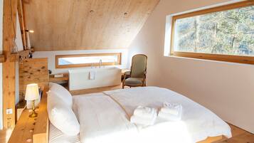 Une chambre avec un plafond en bois, une grande fenêtre, un lit, une chaise, une table de chevet avec une lampe et une petite commode en bois.