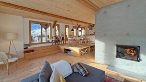A spacious living area with a fireplace, wooden flooring, and large windows offering a view of the outdoors. - Design chalet with Alpine panorama (Riom)