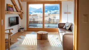 A cozy living room with a wooden ceiling, a large window with a view of mountains, a flat-screen TV, a wooden coffee table, and a sofa with cushions. - Design chalet with Alpine panorama (Riom)