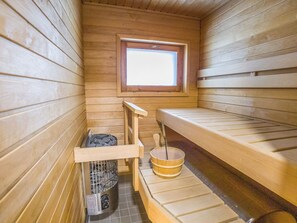 A sauna room with wooden benches, a window, a bucket, and a stone-filled container. - Operomukka 5 by Interhome (Kittilä)