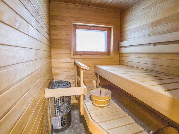 A sauna room with wooden benches, a window, a bucket, and a stone-filled container.