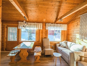 A cozy cabin interior with a wooden ceiling, a sofa, a chair, a table, and two windows with curtains. - Operomukka 5 by Interhome (Kittilä)