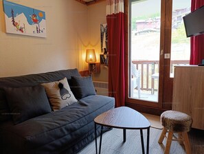 Un salon confortable avec un canapé, une petite table et un tabouret. Il y a une fenêtre avec des rideaux, une lampe fixée au mur et un tableau.