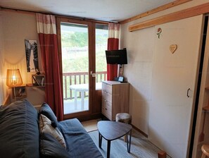 Quarto mobiliado com sofá, uma mesa pequena, televisão e porta de vidro deslizante que dá para uma área externa.