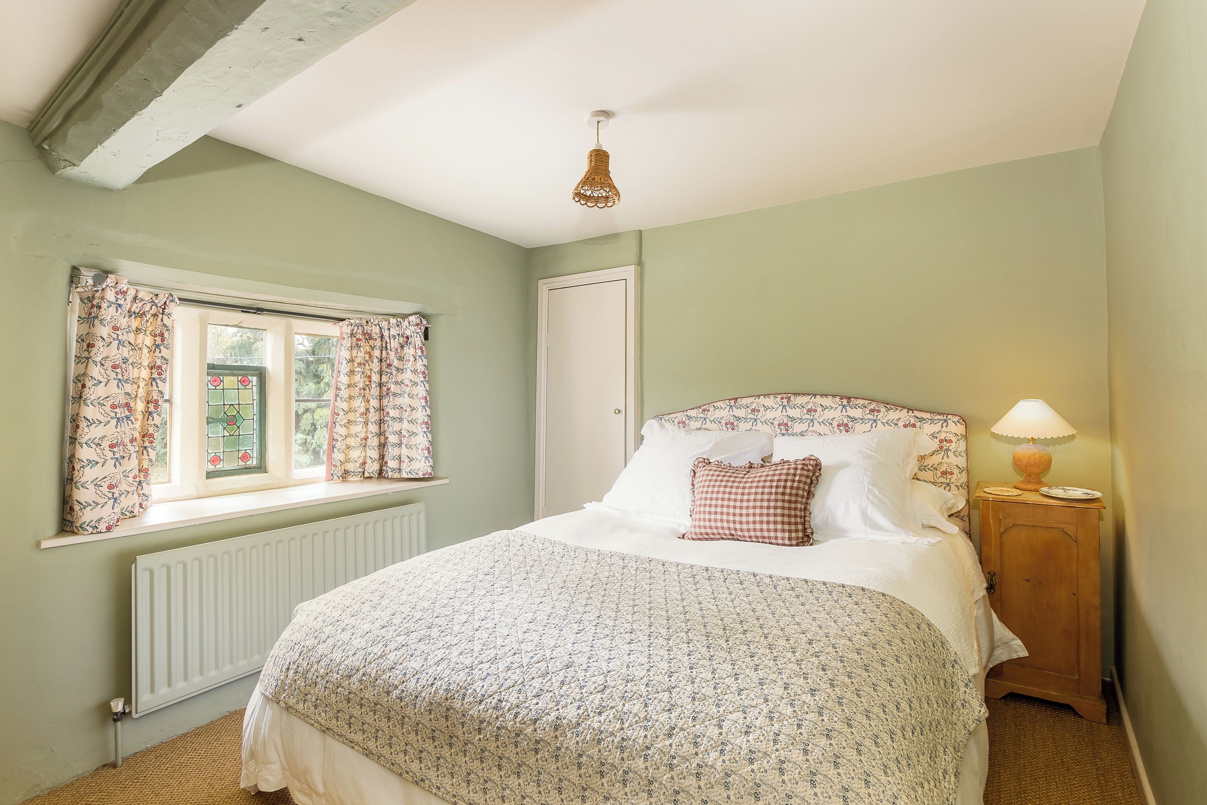 A bedroom with a bed, window with curtains, radiator, wooden nightstand with a lamp, and a door.