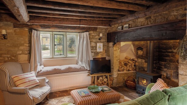 A cozy room with a stone fireplace, a television on a wooden stand, a cushioned bench, and a window with curtains. - Charming 2BD in the Heart of Kingham (Chipping Norton)