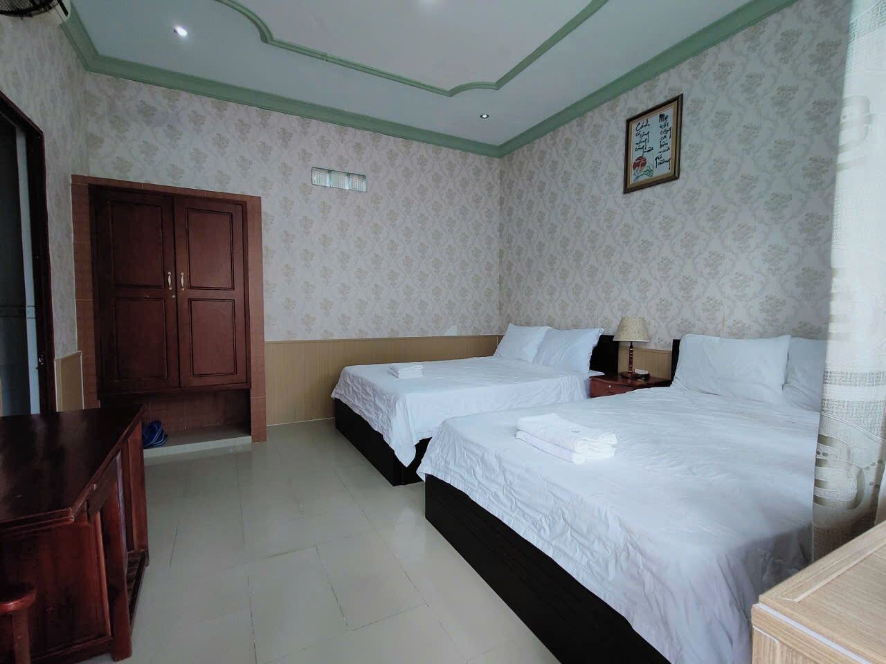 A hotel room with two beds, a wardrobe, a desk, and a small table with a lamp.
