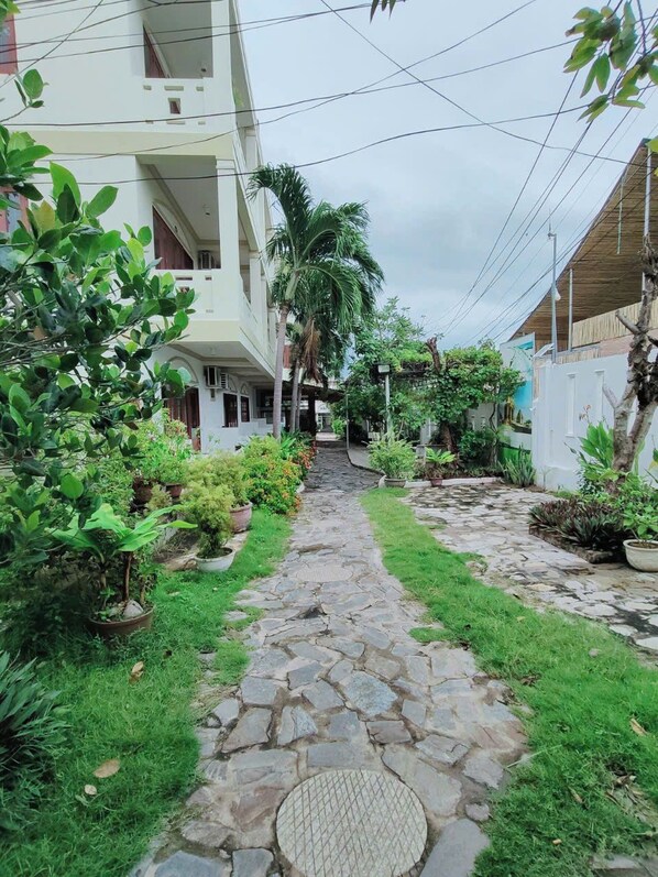 A stone pathway leading to a building with a balcony, surrounded by lush greenery and potted plants. - Min's Homestay (Mui Ne)