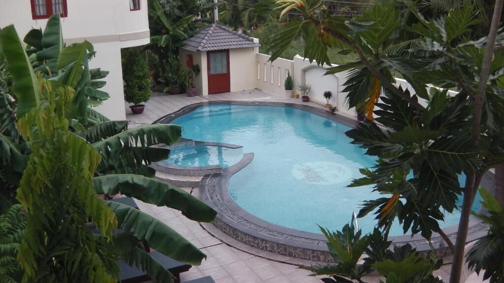 Una piscina rodeada de plantas tropicales y un pequeño edificio con una puerta roja.