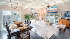 A spacious living area with a dining table set for four, a white sofa, and a television mounted on the wall.