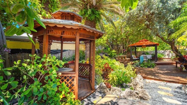 A wooden gazebo with a lattice roof, surrounded by lush greenery and a stone pathway.