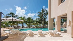 Un área junto a la piscina con sillones de descanso y sombrillas, palmeras y una vista despejada del océano.