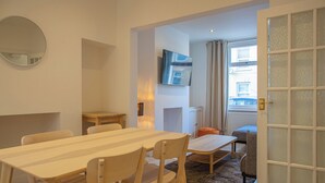 A modern living room with a wooden dining table, chairs, a TV mounted on the wall, and a view of the outside through a window with curtains. - Bright & Airy 2BD - 10min walk to Centre (Cheltenham)